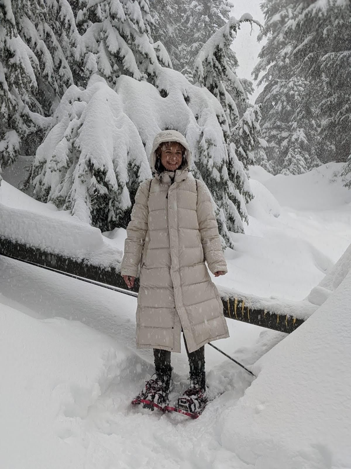 A woman in a white poofy jacket and snowshoes smiles as she poses on a snow-covered trail through tall pine trees.