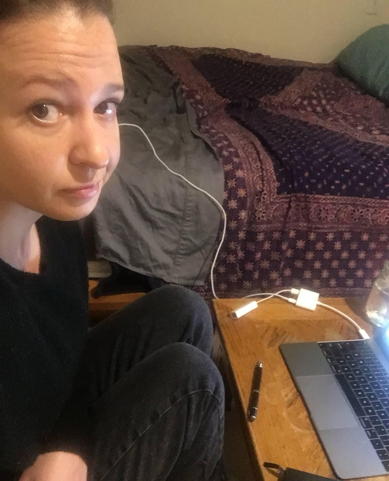 A woman poses for a selfie while sitting next to her laptop and bed.