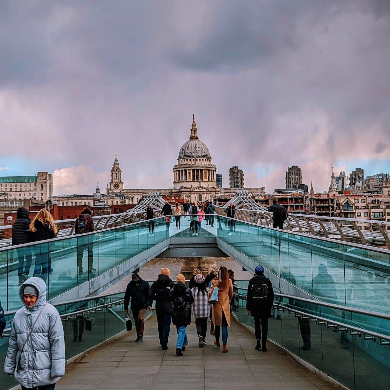 Crowds of people standing on a glass bridge leading to a large domed church.