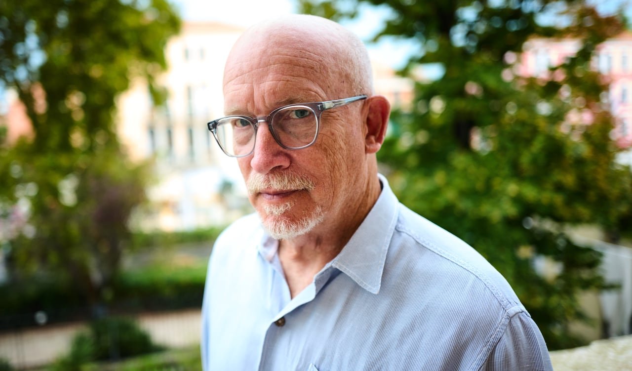 default-180 A bald man with glasses and a white goatee looks into the camera in a shoulder-up photo.